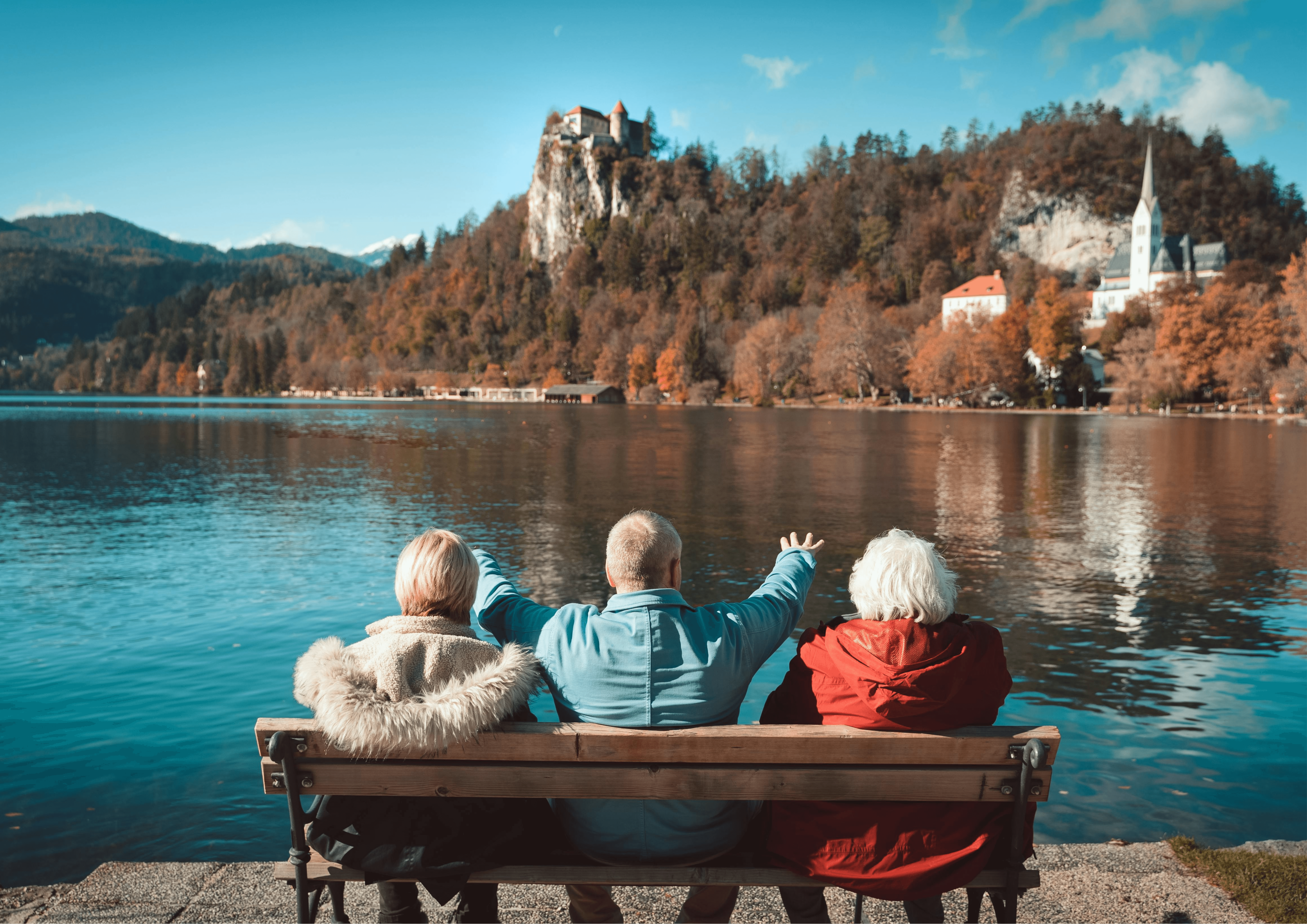 Drei Senior*innen sitzen auf einer Bank an einem See, das Foto ist von hinten aufgenommen.