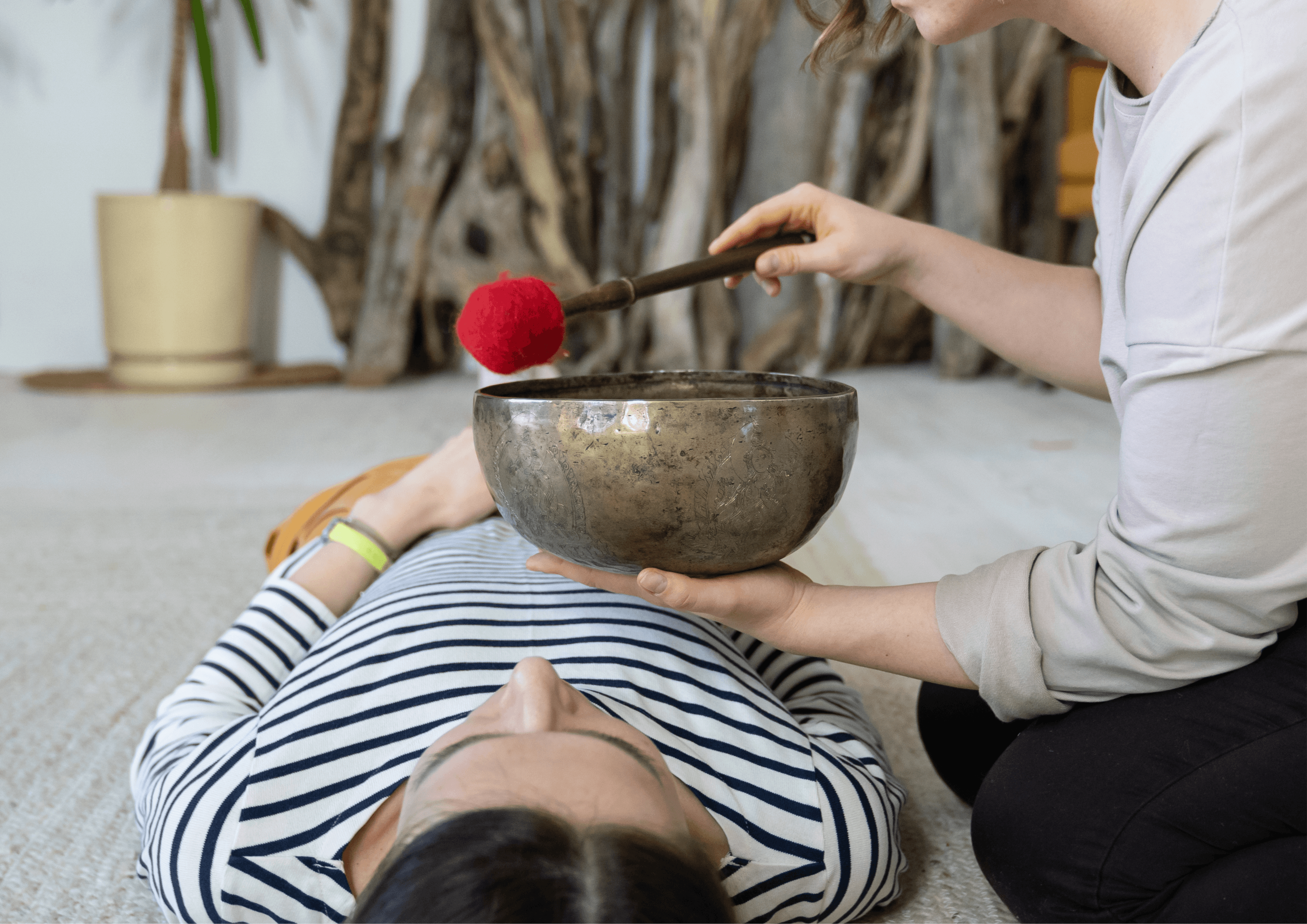 Frau in gestreiftem Oberteil liegt am Boden, jemand hält eine Klangschale über ihren Körper.