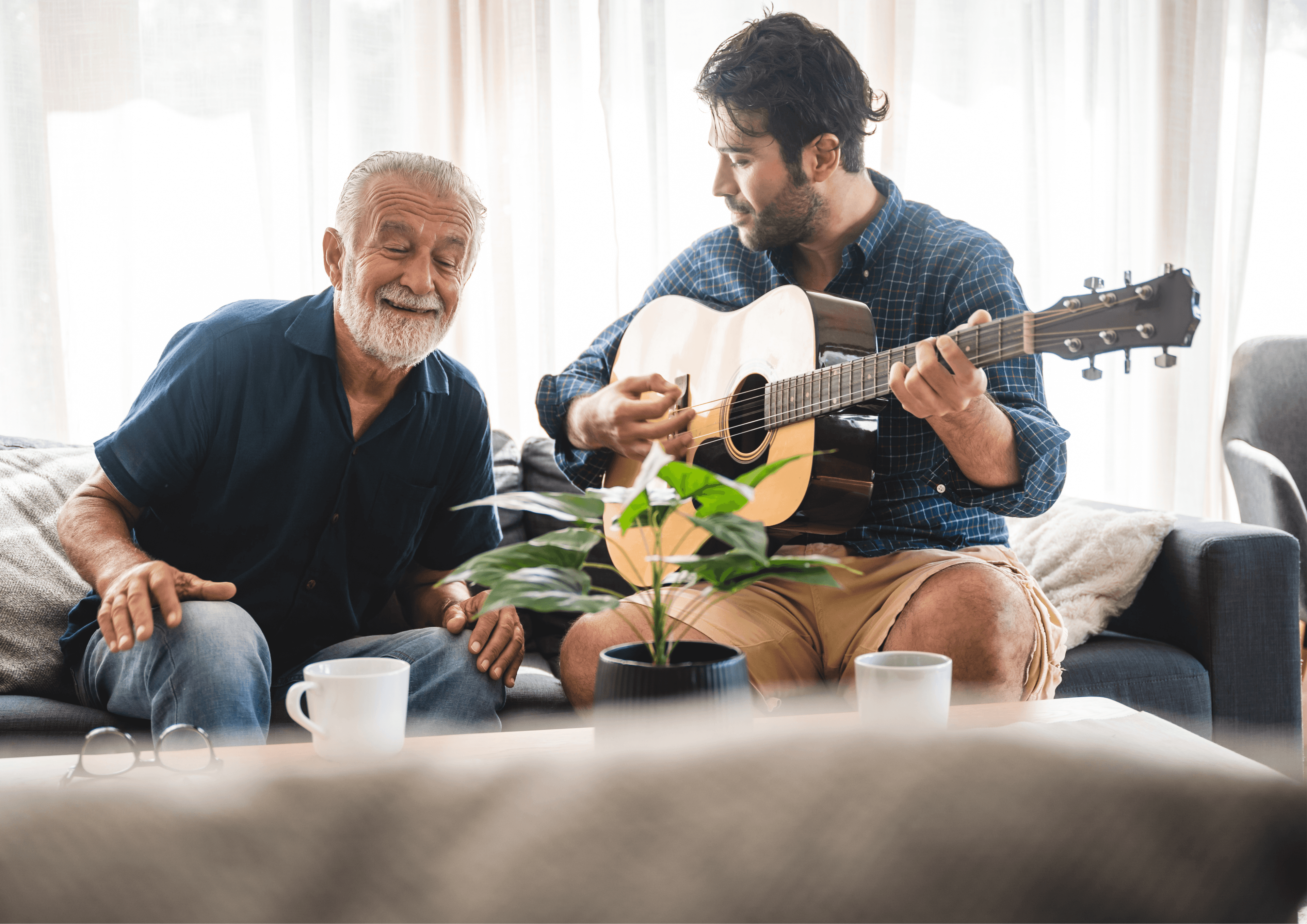 Ein älterer und ein jüngerer Herr sitzen auf einem Sofa, der jüngere spielt Gitarre, der ältere lächelt und hört zu.